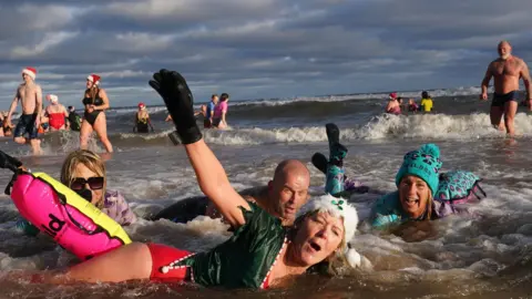 Medios de comunicación de PA: Cuatro personas yacen boca abajo en aguas poco profundas. La mujer del frente lleva un disfraz de elfo con gorro de Papá Noel y levanta un brazo. Las personas detrás de ella sonríen y ríen. A lo lejos, la gente sale del mar.