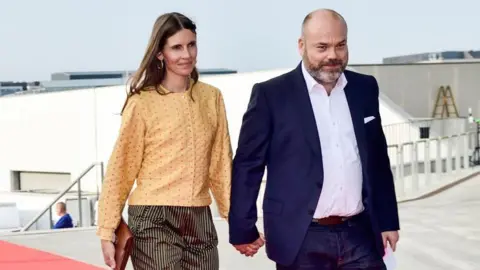 AFP Anders Holch Povlsen wearing a navy suit, holding his wife's hand. She is wearing an orange cardigan and striped trousers.