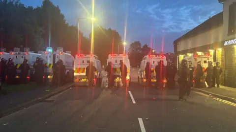 BBC A line of police in riot gear. It's night. A number of police land rovers are parked. 