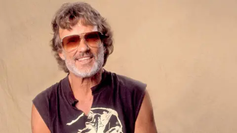 Kris Kristofferson poses for a photograph in the 1970s against a beige background, wearing sunglasses and a black sleeveless t-shirt