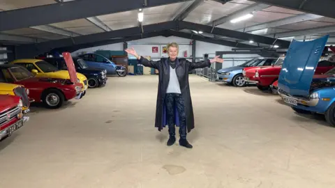 A woman with short blonde hair and wearing a long black leather coat stands in the middle of two rows of classic cars, holding her arms up to point at them.