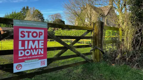 A red sign with white writing urging people to "Stop Lime Down" is fixed on a five bar wooden gate. Behind the gate is a grassy field, and a stone house.