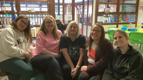 Five staff members at Stoke Primary School sit on a sofa in the classroom of their alternative provision The Aviary. Behind them is a green classroom table and chairs and doors to an outdoor play area