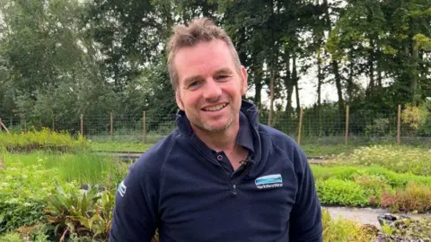 Mr Hampshire is wearing a blue quarter zip fleece with a Yorkshire Water logo on the left breast pocket. He is smiling and standing in front of a river bank with a variety of plants growing alongside.