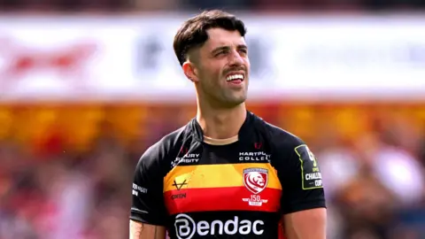 Rugby's Adam Hastings wearing black t-shirt with two stripes in red and yellow. He has brown hair and is frowning.