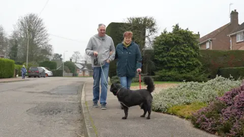 Jamie Niblock/BBC Lynne and John walk their black, three-legged dog Max. Lynne is clutching a red walking stick in her left hand, while holding John's arm with her weakened right- hand.