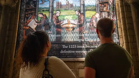 Durham Cathedral A man and woman looking at a stain glass window at the cathedral. A message on the window states the cathedral holds the only copy of the 1216 Magna Carta. The scene on the window shows four monks writing on paper. 