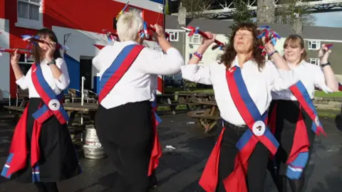 Five people clog dancing outside in the sunshine. They are all wearing white shirts with black trousers and red and blue sash's. 