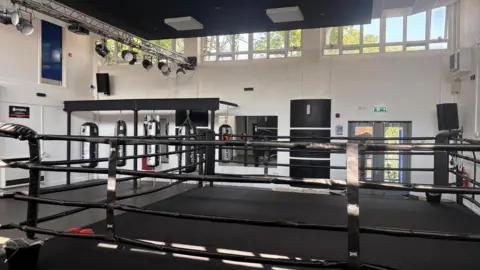 Supplied A spacious boxing gym with a large black boxing ring in the foreground. Several heavy punching bags hang in the background beneath a metal lighting rig fitted with stage lights.