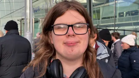 Vikki Irwin/BBC A young man is standing outside a council building with protesters in the background. He has long hair, wears glasses and is smiling. He has headphones around his neck. 