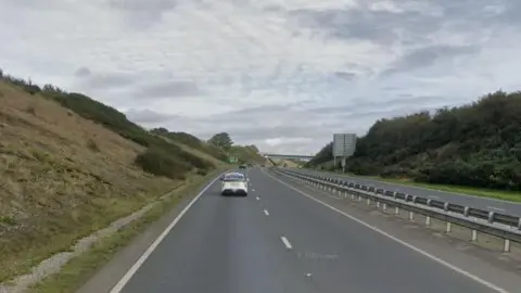 A white car drives along the A38 between Dobwalls and Liskeard in Cornwall on a cloudy day. Large grass verges with bushes line both sides of the road. Several other vehicles are further ahead on the road in the distance. A bridge is also in the distance.