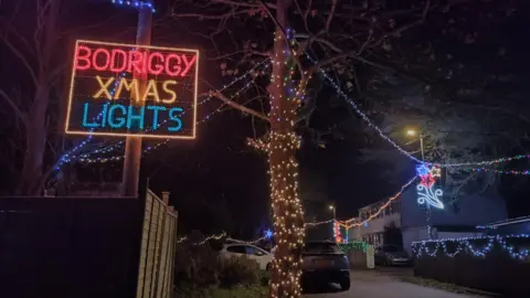 A Christmas light display which states Bodriggy Xmas Lights. A tree next to the sign is covered in warm white Christmas fairy lights. Multicoloured lights are lining the street behind.