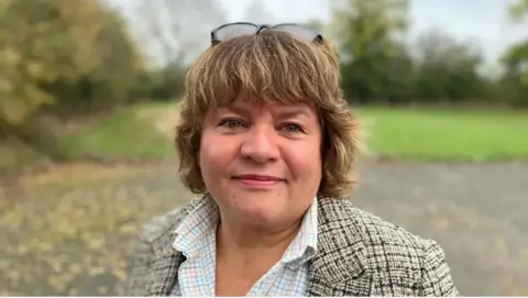 A close-up of Nicola Gribble with short light-brown hair, wearing a patterned jacket and collared shirt, with glasses resting on top of the head. She stands outdoors on a paved path, with grassy areas and trees in the background under soft natural light.