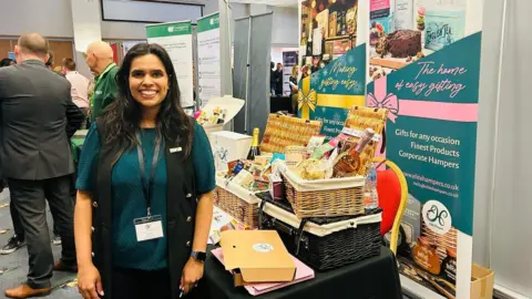 Handout Jeevan Punj stands at a stall displaying her hampers at a business event. She is wearing a green top and has a lanyard. Hampers containing food and drink are displayed on a table next to her. There are signs with information about her company.