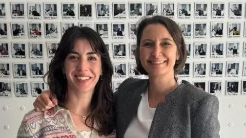 Susannah de Jager Two women with brown hair smile at the camera. The woman on the right has her arm around the woman on the left. Dozens of Polaroid photos are stuck on the wall behind them.