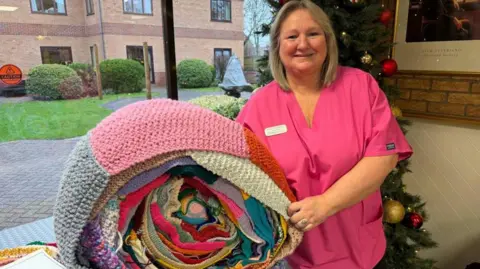 Activities coordinator Caroline Hughes pictured next to the rolled up multi-coloured giant scarf. She is wearing a pink top and is smiling at the camera. There is a Christmas tree with baubles in the background.