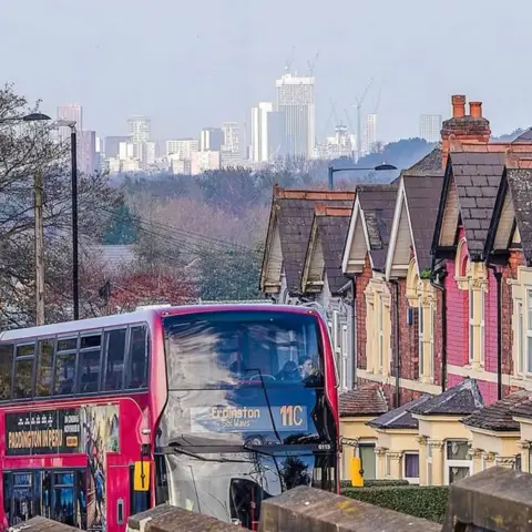 Damien Walmsley A red bus, saying 11C, travels past a row of terraced houses with a skyline of Birmingham city centre in the background. 