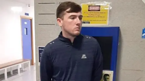 Jack Kavanagh looking away from the camera in a photo taken by the National Crime Agency.  He has short brown hair and is wearing a dark blue sports top with a zip at the neck.