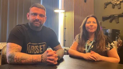 A man and woman sat at a table, both looking at the camera and smiling. The woman wears a floral top, the man a t-shirt with the words Operation Reset on it
