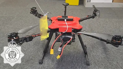 A drone with a red-coloured body and black arms, legs and propellers. It has been placed on the grey-carpeted floor of an office.
