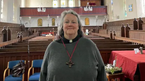 A person with a grey sweater and dog collar with a cross hanging on the chest with a church and pews in the background.