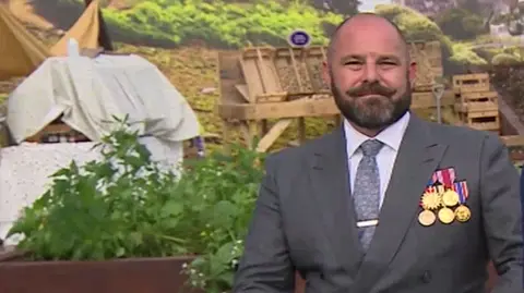 BBC James Kaye, who has a beard and is in a grey suit decorated with five medals