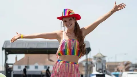 Amy LT Photography A woman wearing a rainbow top, skirt, and hat with her hands in the air