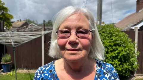 Judy Washington poses for the camera in her sunny back garden. She has glasses and short, straight white hair and wearing a blue patterned dress. A clothing line, bush and garden fence can be seen in the background.