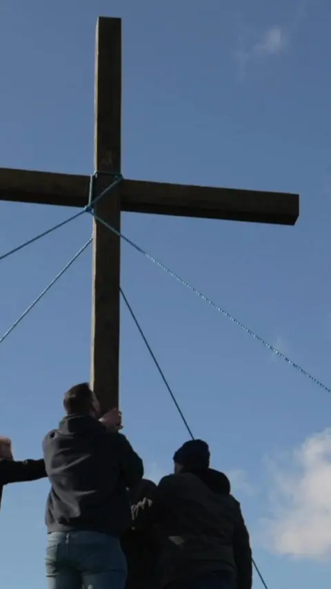 A wooden cross is put up on a hill. Three people's silhouettes are in the foreground.