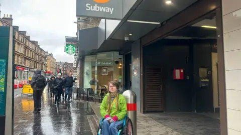 BBC A wheelchair user in a green coat and blue jeans, sat outside a Subway station 