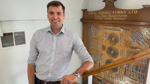 BBC / Jack Hadaway-Weller A man in a white checked shirt leans on a bannister in front of a display case full of tuning forks. He is smiling and wearing a silver watch.