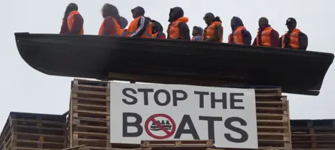 PA Media The picture shows a model of a small boat with figures inside in orange fluorescent vests. Below is a sign which says 'STOP THE BOATS'