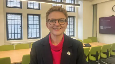 Photograph of Bev Craig, the leader of Manchester City Council. She has short brown hair and is wearing a red roll-neck top with a black blazer and dark nrown glasses. She is smiling. The image shows her inside her office at Manchester Town Hall with a large boardroom table with about a dozen lime-green coloured chairs.