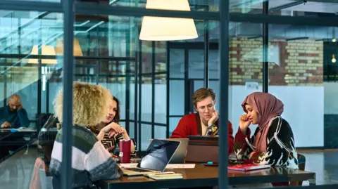 10'Al hour/Getti three young women and a young man sitting around a table with laptops in a glass meetings room in the office.