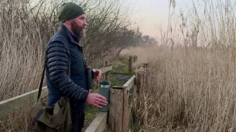 Paul Hadaway, a tall dark bearded man, looks out over a fence across a reedbed. He has a satchel and binoculars.