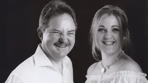 A black and white image of a father and daughter. The father is wearing a white shirt. The daughter is wearing a white top has a chain necklace.