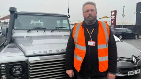 A man in a high vis jacket, with grey hair and beard standing infront of a landcover, at a service station