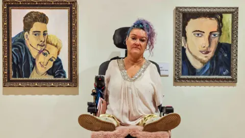 Alamy Alison Lapper sits in her wheelchair besides two portraits she painted of her son Parys. The portrait on the left is a painting of Parys cuddling Alison. They are smiling. The portrait on the right is only of Parys and his eyes are widened with a pensive look on his face. 