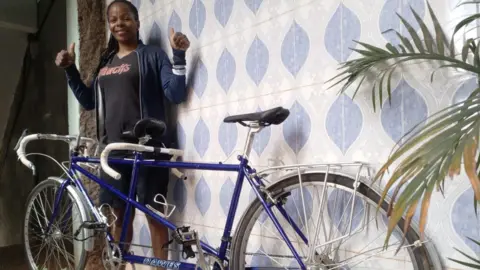 A woman wearing a black T-shirt and blue jacket smiles and holds two thumbs up while standing behind a blue tinted tandem bike