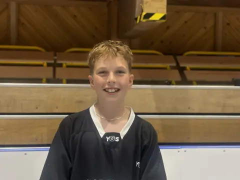 BBC News Archie is a young, fair-skinned boy with blonde hair wearing a black jersey with white on the neck. He is standing in the roller rink with a side wall behind him.