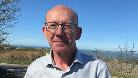 BBC A man in an open neck light shirt, with a bald head and wearing glasses