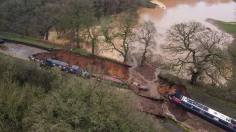 PA Media A large muddy hole, seen from the air, with two canal boats in it and a large brown pool of water at the top of the image