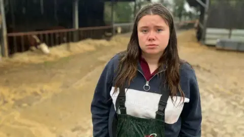 Maria Warne-Elston stands in a yard with cattle sheds in the background. There are cows and young calves eating hay in the sheds.