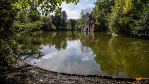 A scenic view of the Roman Boathouse within Birkenhead Park, with overhanging tree branches in the foreground and a lake.