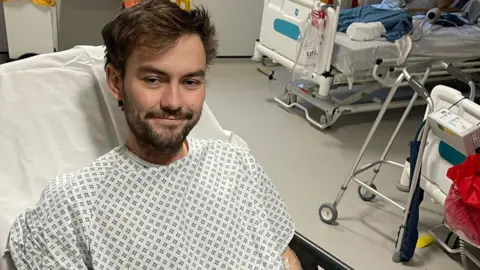 Michael Adams Michael in a hospital gown and bed looking thin and unwell, but still smilling