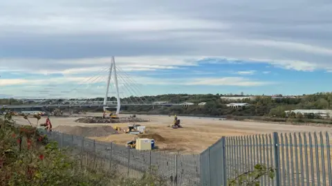 A building site covered in sand with a few construction vehicles on it, with grey fencing and greenery in front. There are a few piles of rubble on the ground. In the background is the Northern Spire bridge, which has a very tall white pointed arch in its centre with cables shooting off. Other buildings and trees are behind it. It is a cloudy day but blue skies can be seen in the background.