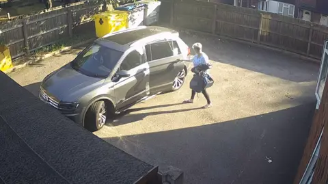 Dudley Metropolitan Borough Council Jamiel Slaney-Summers getting into a grey car. She is shutting the door. She is wearing a white top with butterflies on. She has short, grey hair. She looks like she is in a courtyard somewhere. We can see some bins in the background. 