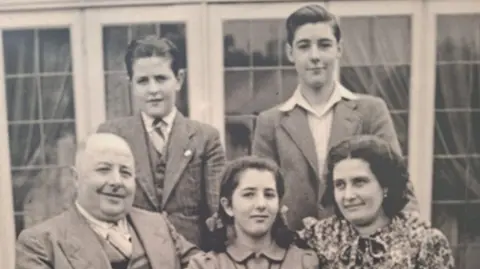 Family photo A black and white photo of a family. Eddie Rabaiotti stands on the left behind his dad, sister Louisa and mum, and next to brother Don. The boys wear smart suits and Louisa has her hair in pigtails and is wearing a collared dress. Her mum is sat next to her wearing a floral dress which ties at the neck. Dad sits on the left wearing a suit, vest and white shirt. 