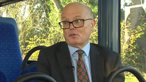 Andrew Wickham sits on a Bluestar bus, the seats are blue with pale blue polka dots. He wears thick black framed glasses, and black suit blazer, with a pale blue shirt underneath and a green a pink printed tie.