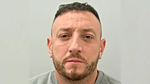 Jake Balmforth is looking straight at the camera in this police mugshot. He has short brown hair and stubble and is wearing a grey top.